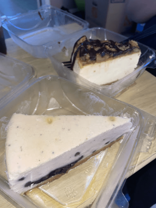 Two slices of cheesecake in plastic containers, one with a chocolate topping and the other with a light pink layer.